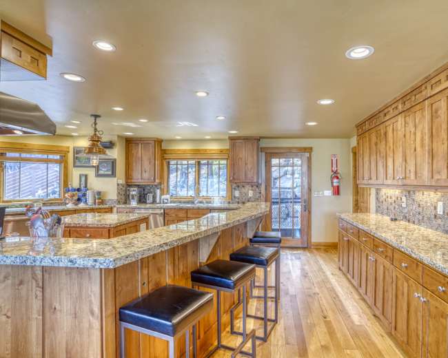 The image shows a spacious kitchen with wooden cabinets, granite countertops, and a central island with black bar stools.