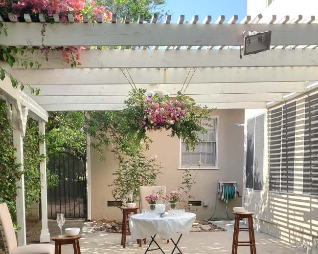 A small patio features a white table set with flowers and surrounded by four chairs and two stools under a pergola with hanging plants.