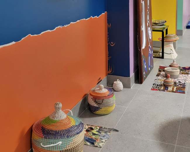 The image shows a colorful interior wall with orange, purple, and blue sections, featuring woven baskets on the floor and a framed artwork hanging above.