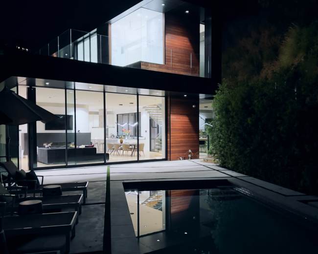 A modern home with large glass windows overlooks a pool, reflecting the structure under a night sky.