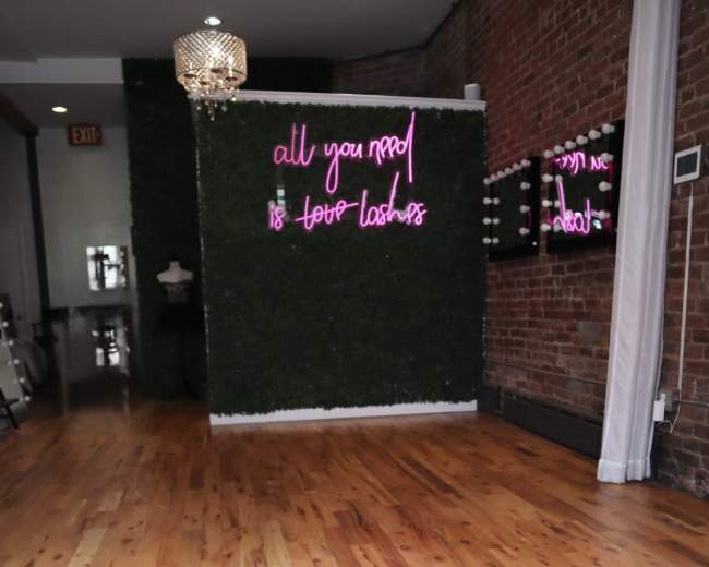 The image shows a room with wooden flooring, a greenery-covered wall featuring neon pink text, and a vanity area with mirrors and lights.