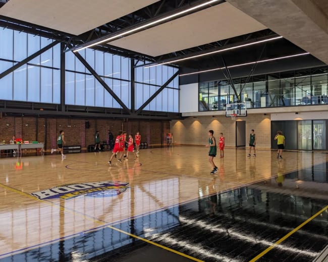 A basketball court features players in red and green jerseys engaging in a game, with large windows allowing natural light into the space.