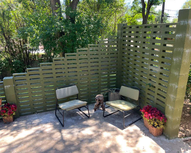 A small outdoor seating area features two metal chairs, a rustic side table, and potted flowers against a vertically slatted green wall.