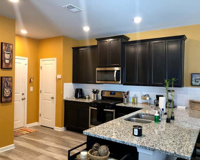 The image shows a modern kitchen with dark cabinetry, a granite countertop, and stainless steel appliances, illuminated by overhead lights.