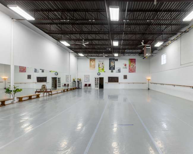 The image shows a spacious, empty dance studio with a mirrored wall, ballet barre, and bright lighting.