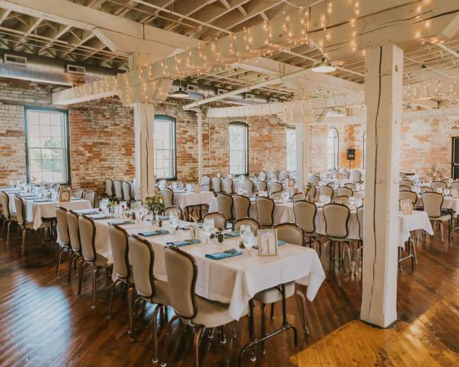 A large banquet hall features multiple long tables set with white tablecloths, surrounded by chairs, against a backdrop of exposed brick walls and string lights overhead.