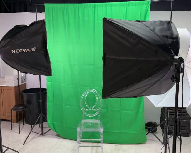 The image shows a photography setup with two softbox lights and a clear acrylic chair positioned in front of a green backdrop.