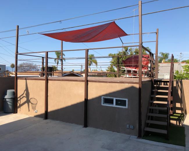 The image shows a rooftop area with a red shade sail, surrounded by metal railings, and accessed by a staircase on one side.