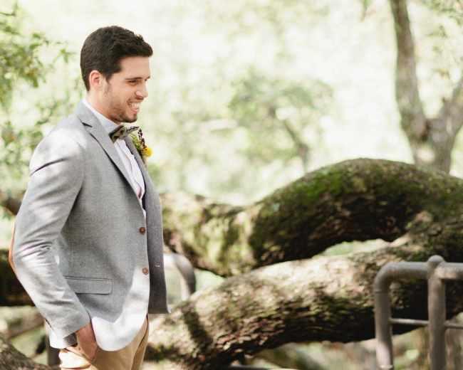 A man in a gray jacket stands near a large tree trunk while smiling at a woman out of frame.