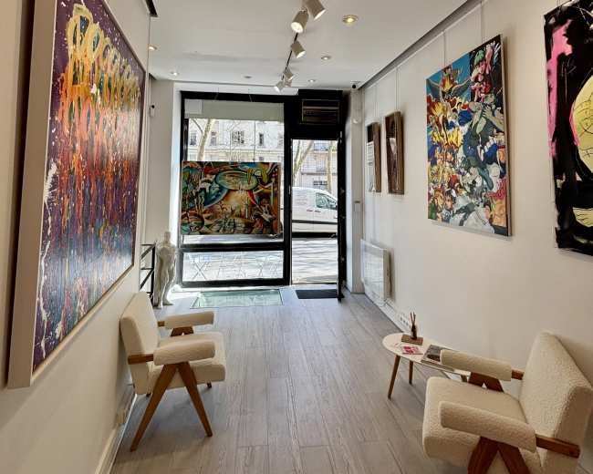 The image shows an art gallery with colorful paintings on the walls and two white chairs positioned near the entrance.