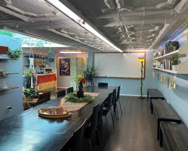 The image shows a spacious classroom or meeting area with a long black table, several chairs, a whiteboard, and colorful decorative elements, including plants and lighting.