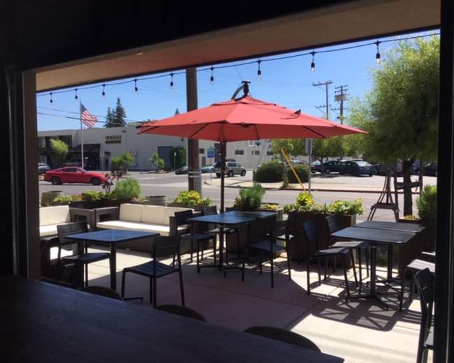 A view from inside a cafe shows a patio with tables and an umbrella, overlooking a street with a flag and parked cars.