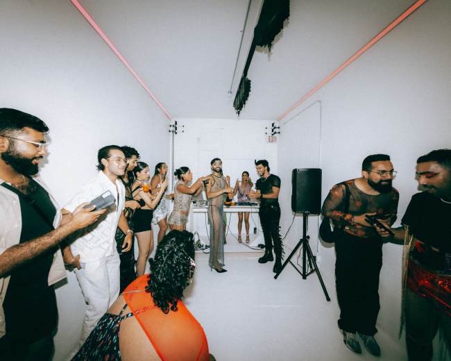 A group of people interacts in a white room with a DJ setup and colorful lighting, while one person stands at a microphone.