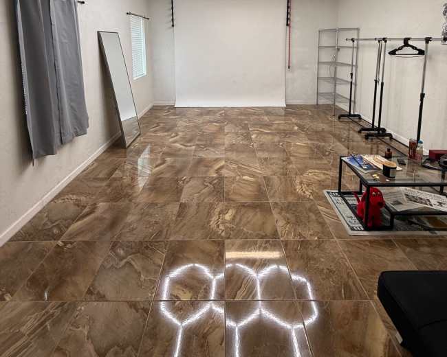 The image shows an empty room with glossy tiled floors, a light-colored wall, a hexagonal ceiling light design, a large mirror, and some clothing racks along with a backdrop setup.