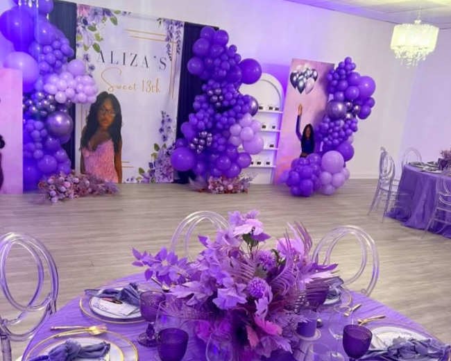 A decorated event space for a sweet 18th birthday celebration, featuring purple-themed decorations, table settings, and a backdrop with balloons and a portrait.