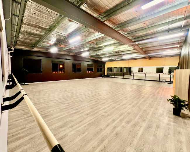 The image shows a spacious, well-lit dance studio with a mirrored wall, wooden flooring, and barre fixtures along one side.