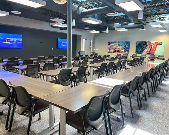 A spacious meeting room with multiple rows of tables and chairs, two screens mounted on the front wall, and large windows allowing natural light.