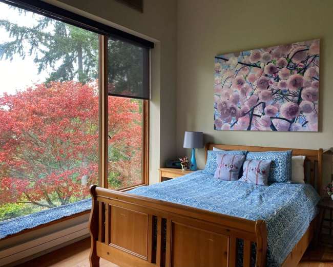 A bedroom features a wooden bed with a blue patterned bedspread, decorated with pillows, and a large window that shows a vibrant red tree outside.