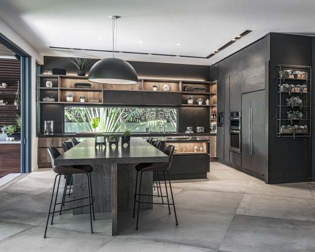 A modern kitchen features dark cabinetry, a large dining table, and a window displaying greenery outside.