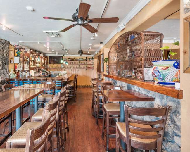 The image shows a restaurant interior featuring wooden tables and chairs, a bar area with shelves of liquor, and a stone wall accent.