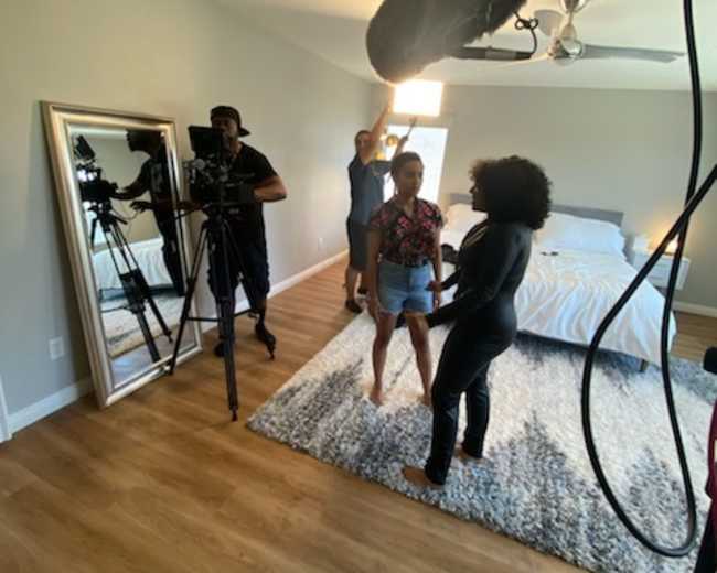 A group of three people is filming a scene in a well-lit bedroom with a large bed and a mirror.