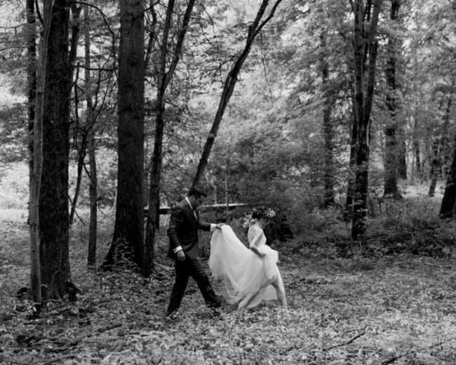 A man in a suit walks with a woman in a wedding dress through a forested area.