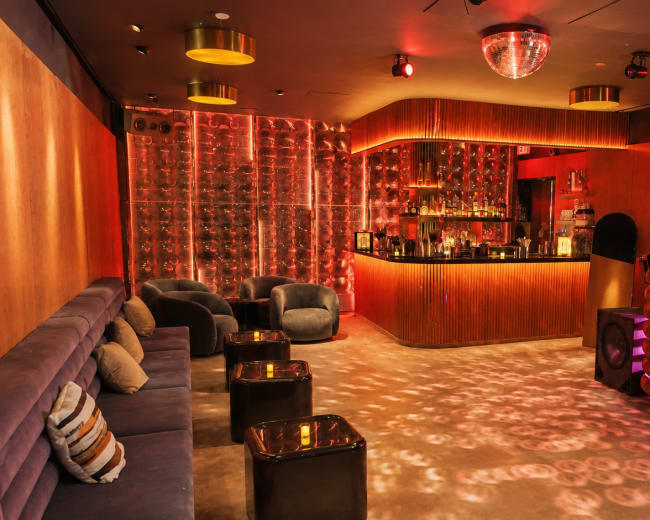 The image shows a stylish bar area with a circular counter, plush seating, and decorative lighting, creating a vibrant nightlife atmosphere.