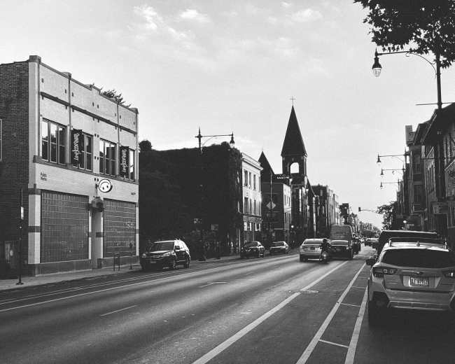 A wide street with vehicles parked along the sides, featuring a mix of buildings, including a church with a tall spire in the background.