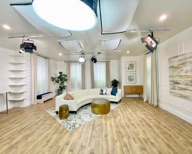 A well-lit studio space featuring a curved white sofa, a round gold table, and decorative plants against a backdrop of wall art and curtains.