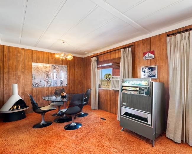 A retro-styled living room with wooden paneling, a glass dining table, and a vintage beverage cooler against a backdrop of desert scenery visible through large windows.