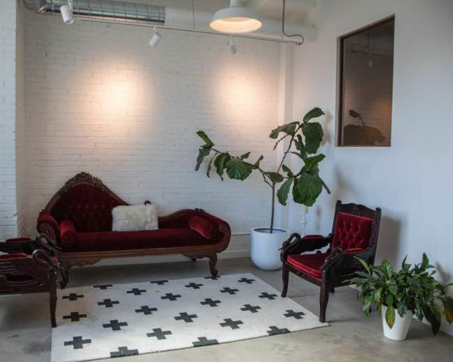 The image shows a seating area with a red velvet sofa, two matching armchairs, a potted plant, and geometric-patterned rug against a white-brick wall.