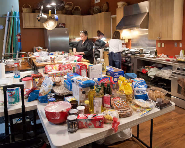 A busy kitchen scene features two women cooking while a table is cluttered with various food items and ingredients.