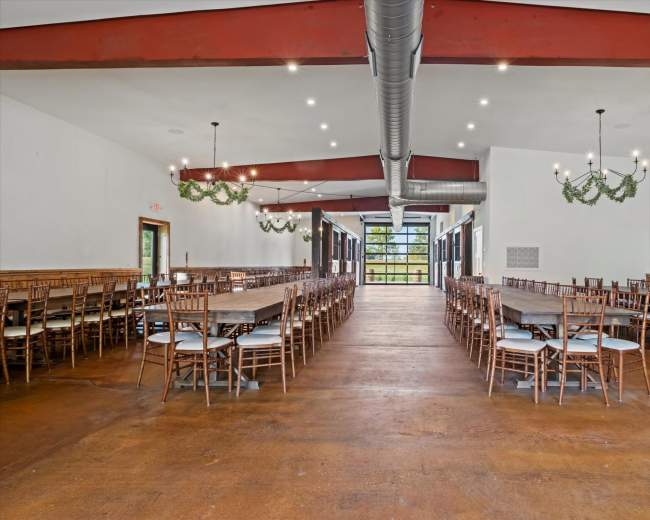 The image shows a spacious event hall with rows of wooden tables and chairs arranged in two parallel lines, flanked by large windows and decorative lighting fixtures.