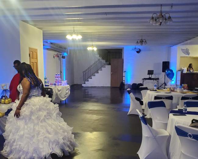 A woman in a white bridal gown stands in a decorated hall with tables covered in white and blue, prepared for a celebration.