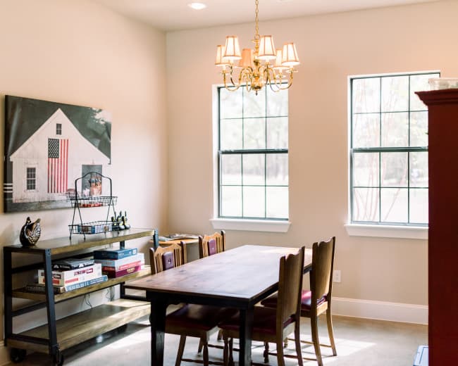 A dining area features a wooden table with chairs, a chandelier overhead, large windows, and a wall displaying a framed image of a house.