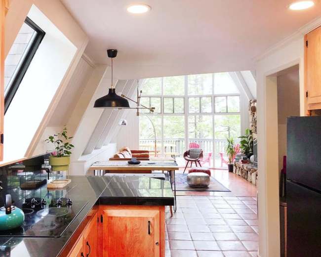 A kitchen with wooden cabinets and a black countertop opens into a sunlit dining area featuring large windows and a rustic table.
