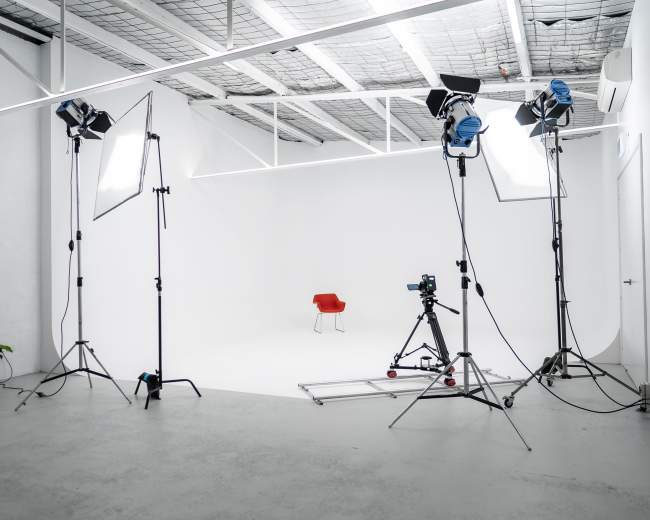 A minimalistic photo studio features a red chair positioned at the center, surrounded by lighting equipment and a camera on a track.