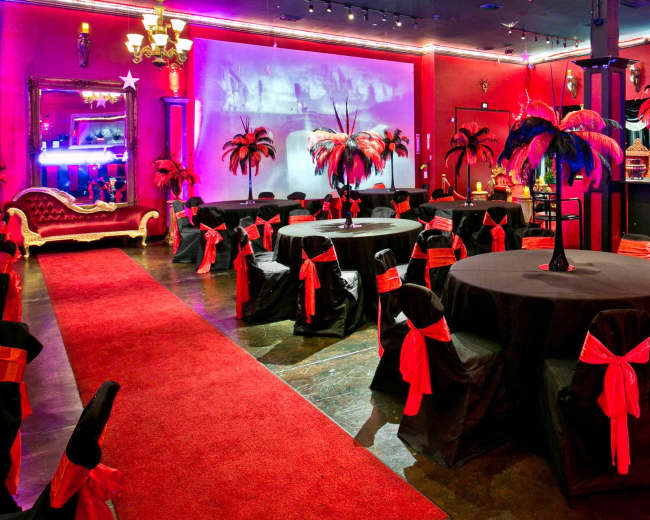The scene features a vibrant event space with black tables adorned with red sashes, black chairs, and decorative palm trees, all set under colorful lighting.