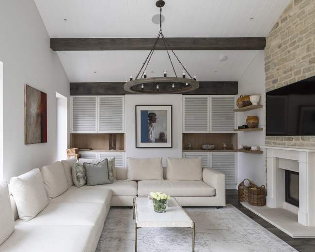 A bright living room features a large light-colored sectional sofa, a round chandelier, and a stone wall with a mounted television.