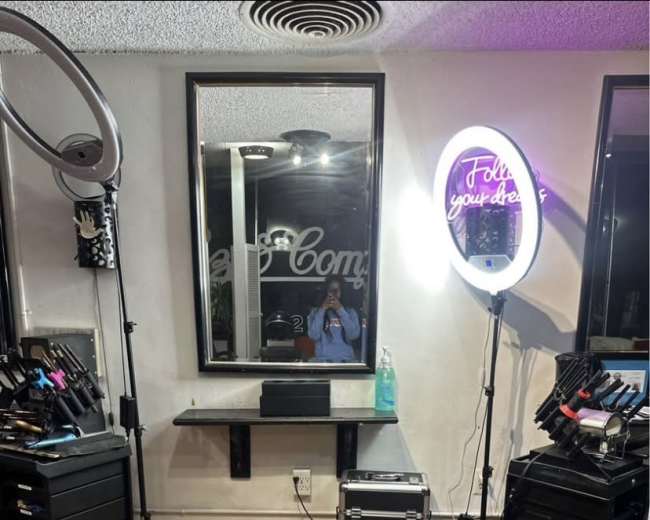 The image shows a hair and makeup studio with a black swivel chair, illuminated mirrors, and various styling tools organized in storage units.