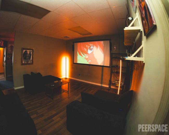 A cozy room features a large projector screen displaying an animated character, with dark couches and a warm light source illuminating the space.