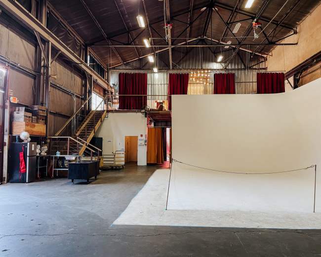 The image shows a spacious industrial studio with a large empty white backdrop, red curtains, and a staircase leading to an upper level.