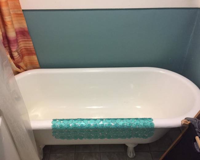 The image shows a white bathtub with decorative blue tiles placed along the edge, situated in a bathroom with a colorful curtain and turquoise wall.