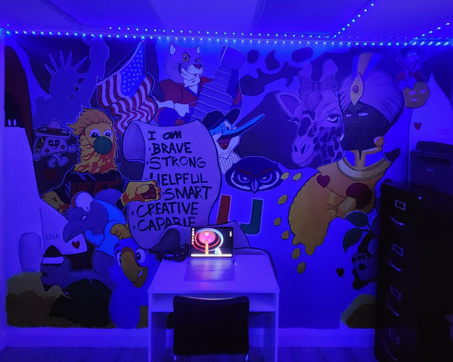 The scene features a brightly colored mural on a wall, depicting various cartoon animals and characters, with a white table and illuminated computer in the center of a dimly lit room.