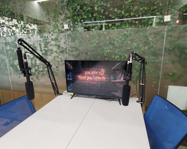 A television displaying a neon sign surrounded by two microphone stands is positioned on a table in a room with greenery on the walls.