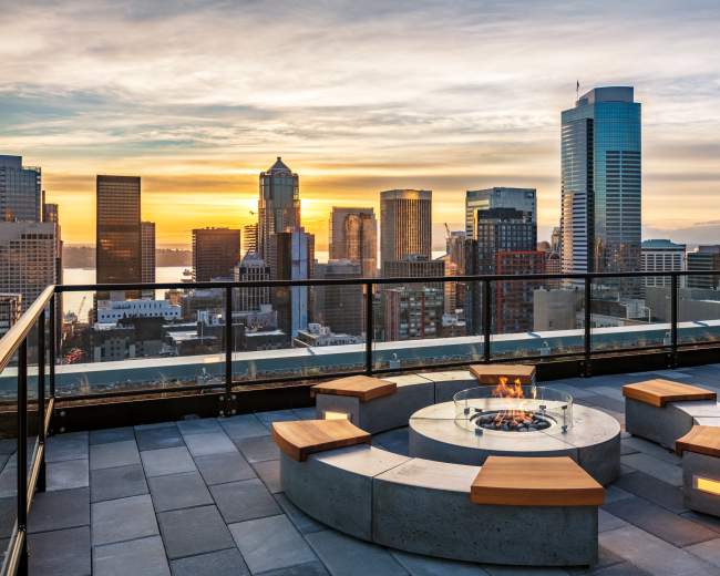 A city skyline at sunset is viewed from a rooftop terrace featuring a circular fire pit surrounded by wooden seating.
