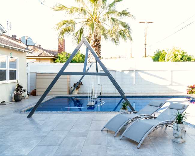 The image shows a backyard pool area featuring an A-frame structure, two lounge chairs, and a palm tree.