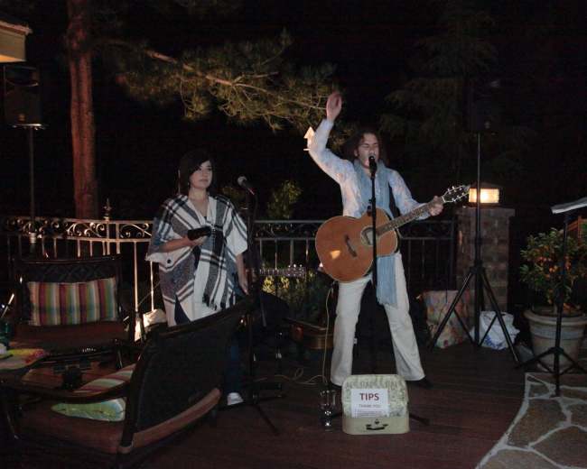Two musicians perform on a deck at night, with one playing guitar and the other standing beside a tips box.
