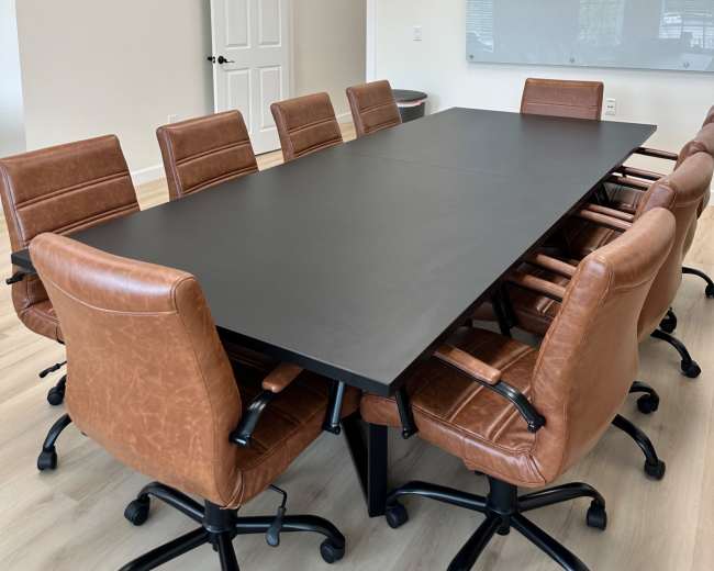 A meeting room features a long black table surrounded by eight brown leather chairs and has a whiteboard on the wall.