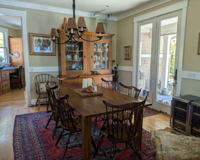 A wooden dining table surrounded by chairs sits in a well-lit room with a china cabinet, rugs, and access to a porch.